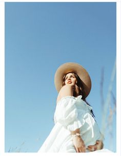 a woman wearing a white dress and hat standing in tall grass looking up at the sky