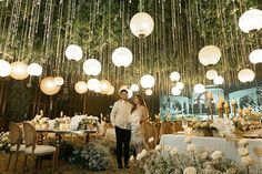 a bride and groom standing in front of a table with lights hanging from the ceiling