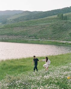 a man and woman holding hands while walking in the grass near a body of water