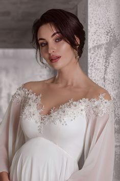 a pregnant woman wearing a white gown with sheer sleeves and an embellishment on her breast