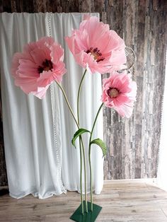 three pink flowers in a green vase on a wooden floor next to a white curtain