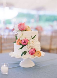 a white wedding cake with pink and orange flowers sitting on top of a table next to a candle