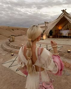 a woman with long blonde hair standing in front of a desert tent holding a pink purse