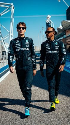 two men in racing suits walk down the street together, both wearing sunglasses and holding hands