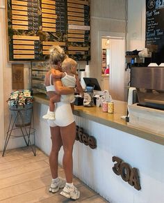 a woman holding a child in front of a counter at a coffee shop with lots of signs on the wall