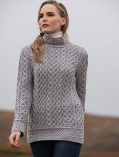 a woman wearing a gray sweater and jeans standing in front of a hill with her hand on her hip