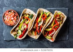 three tacos with meat, vegetables and sauces on a slate platter next to a small bowl of salsa