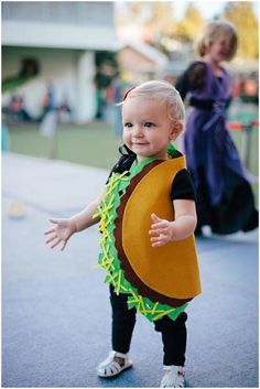 a toddler dressed in a costume standing on the ground with her hands out and smiling