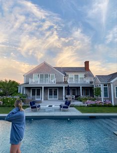 a woman standing in front of a swimming pool next to a large white house with an open porch