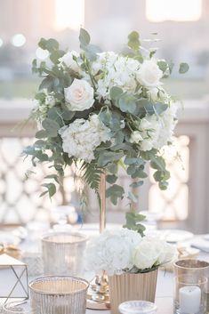 white flowers and greenery sit in gold vases on a table with silver cups