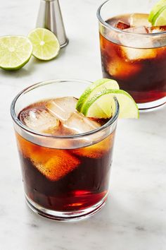 two glasses filled with rum and coke on top of a white marble table next to limes