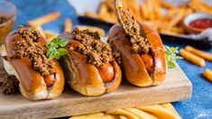 three hot dogs on buns with chili and french fries in the background, ready to be eaten