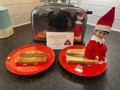 two red plates with sandwiches on them and an elf sitting next to the toaster