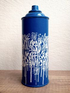 a blue can with white graffiti on it sitting on a wooden table next to a wall