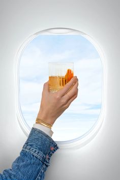 a hand holding a glass with liquid in it and looking out an airplane window at the sky