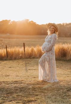 a pregnant woman in a white dress and hat standing in a field with the sun behind her