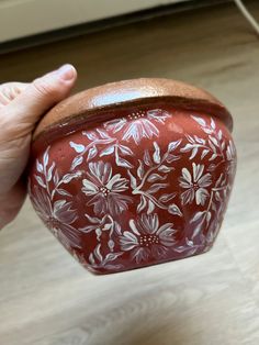 a hand holding a red and white flowered pot on top of a wooden floor