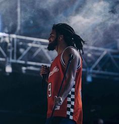 a man with dreadlocks standing in front of a stage holding a microphone and wearing an orange shirt