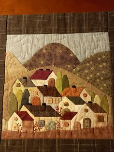 a quilted wall hanging with houses and hills in the background on a wooden table