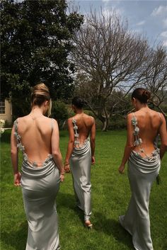 three women in long dresses are walking through the grass with their back to the camera