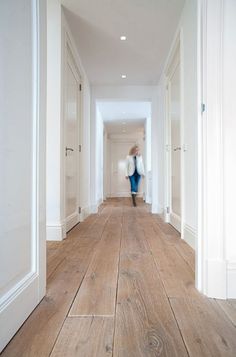 a woman walking down a long hallway with white walls and wood flooring on both sides