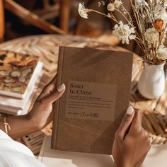 a person holding a book in their hands on a table with flowers and books behind them