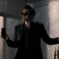 a man in black jacket and sunglasses holding up a cell phone while standing next to a wall