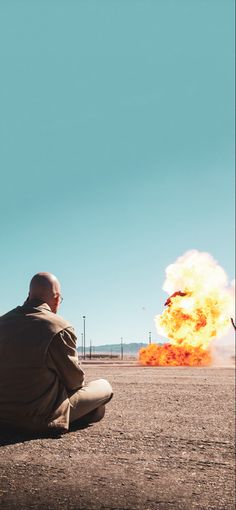 a man sitting on the ground in front of a large fire with flames coming out of it