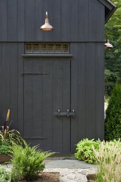 a black shed with a light on the door and plants around it in front of it