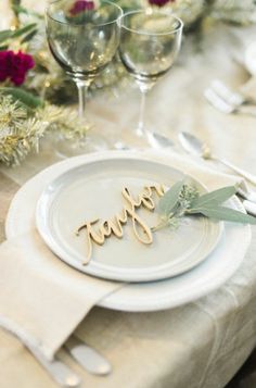 a place setting with wine glasses, silverware and greenery on the table for a festive dinner