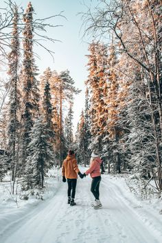 two people holding hands while walking through the snow in front of some tall pine trees