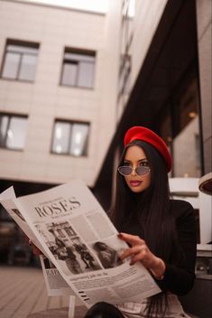 a woman sitting on a bench reading a news paper while wearing glasses and a red hat