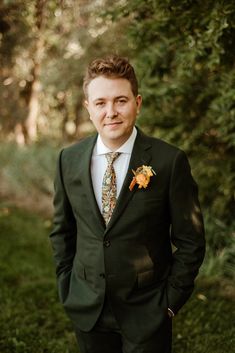 a man wearing a suit and tie standing in front of some trees with his hands in his pockets