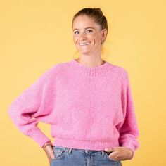 a woman wearing a pink sweater and jeans standing in front of a yellow wall with her hands on her hips