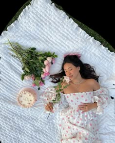 a woman laying on top of a white blanket next to pink flowers and a cake