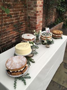 several cakes and pastries are displayed on a long white table with greenery around them