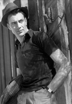 a black and white photo of a man leaning against a fence wearing a fedora