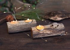 two small candles are sitting on a piece of driftwood with pine cones and lights in the background