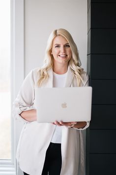 a woman standing in front of a window holding a laptop computer and smiling at the camera