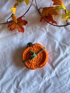 an orange crocheted pumpkin sitting on top of a white sheet next to leaves