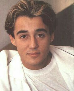 a close up of a person wearing a white shirt and posing for a photo in front of a wall