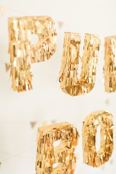 gold foiled letters are hanging from strings on a white wall with confetti streamers