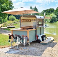 an outdoor food cart with stools and umbrella on the side of it, next to a body of water