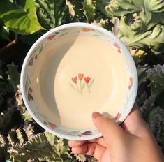 a hand holding a bowl with flowers painted on it in front of some green plants