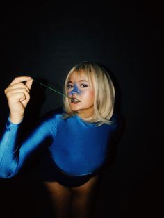 a woman in a blue bodysuit is holding a green string to her mouth and looking at the camera