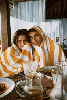 two people wrapped up in blankets sitting at a table with food and drinks on it