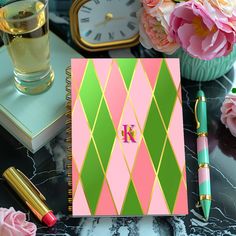 a pink and green notebook with the letter k on it next to flowers, a glass of water and a pen