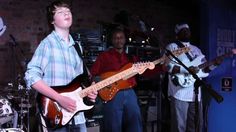 two young men playing guitars and singing into microphones while another man plays guitar in the background