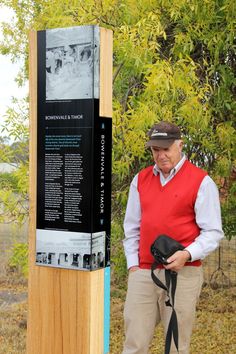 a man standing next to a sign with a camera on it's shoulder and wearing a hat