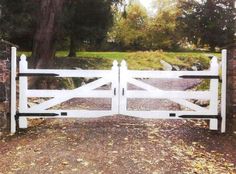 an open white gate in front of a stone wall and trees with leaves on the ground